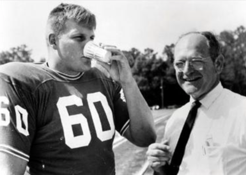 A football player in a jersey drinks from a cup while smiling at a man in a tie beside him on a field.