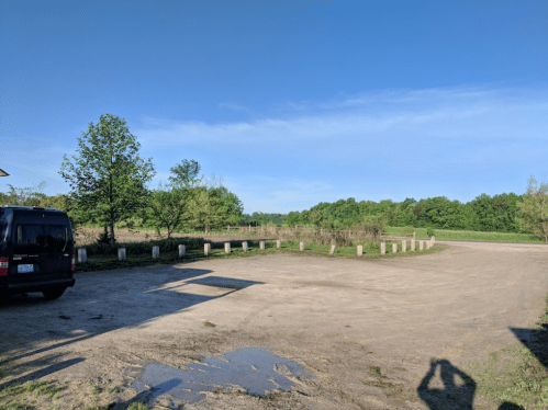 A clear blue sky over a dirt parking area surrounded by trees and grassy fields.