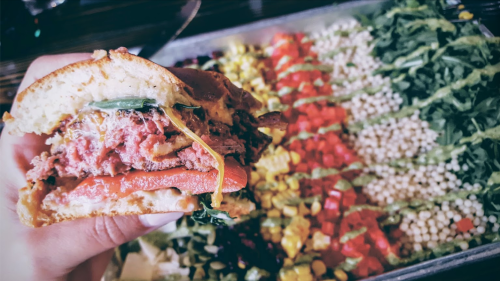 A hand holding a loaded sandwich with various toppings, next to a colorful spread of fresh vegetables and grains.