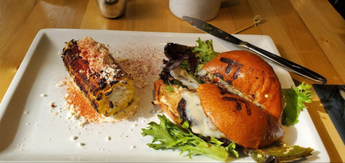 A plate with a grilled burger topped with cheese and lettuce, alongside a charred corn on the cob sprinkled with seasoning.