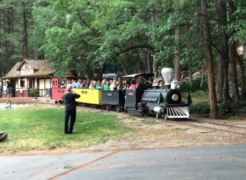 A small train with colorful cars carries passengers through a wooded area, with a station visible in the background.