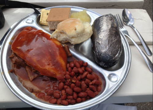 A metal tray with a portion of meat covered in sauce, red beans, a roll, a dessert, and a wrapped item.