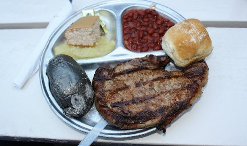 A metal tray with grilled steak, baked potato, red beans, applesauce, cornbread, and a roll.