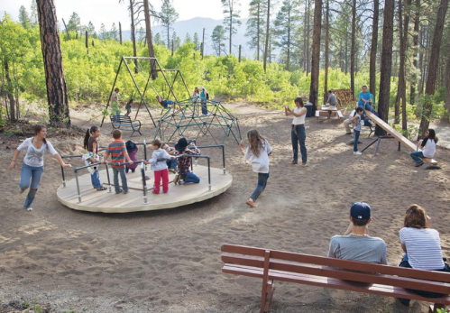 Children play on a playground with a spinning structure and climbing frame, surrounded by trees and mountains.