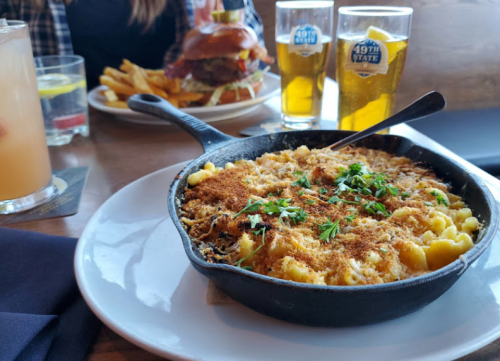 A cast iron skillet filled with baked macaroni and cheese, topped with breadcrumbs and herbs, with drinks and a burger in the background.