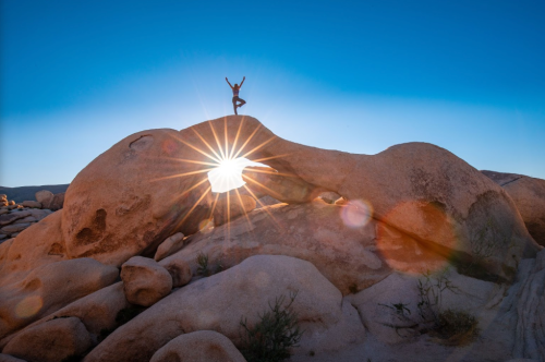 A person in a yoga pose atop a rock arch, with sunlight shining through, creating a starburst effect.