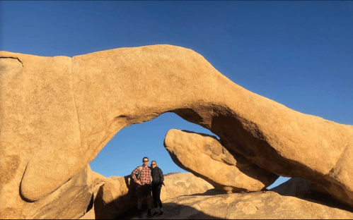 Two people stand under a large rock arch in a desert landscape, with clear blue skies above.
