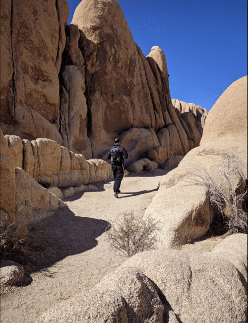 A person walks along a sandy path between large rock formations under a clear blue sky.