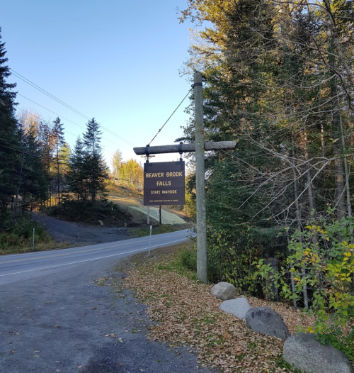 Sign for Beaver Brook Falls State Wayside, surrounded by trees and a gravel road, with autumn leaves on the ground.