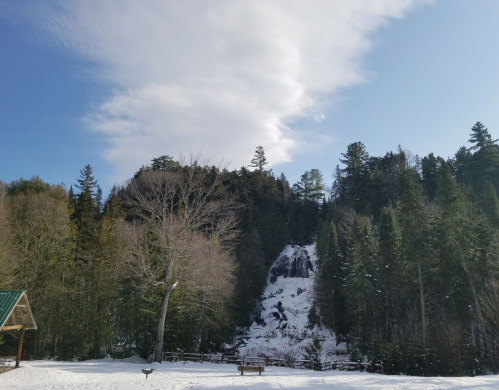 A snowy landscape with a waterfall cascading down a rocky cliff, surrounded by tall trees and a blue sky.