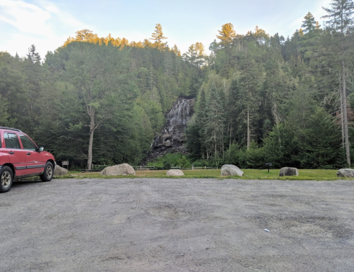 A red SUV parked on a gravel area with a waterfall and dense forest in the background.
