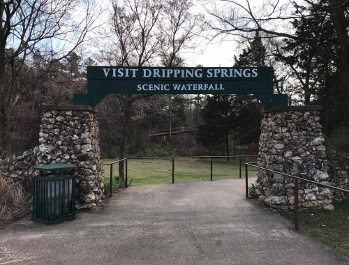 Sign at the entrance to Dripping Springs Scenic Waterfall, framed by stone pillars and surrounded by trees.