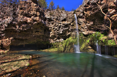 A serene waterfall cascades into a clear pool, surrounded by rocky cliffs and lush greenery under a bright blue sky.