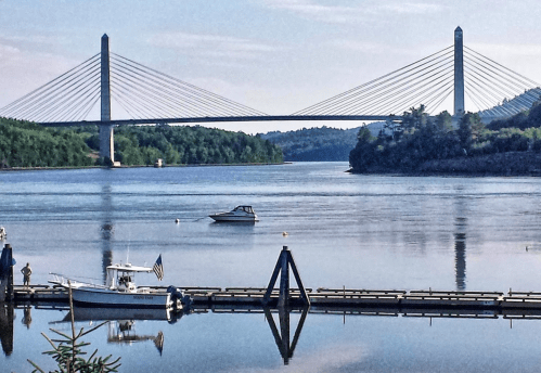 A serene river scene featuring a suspension bridge, a boat on the water, and a dock with an American flag.