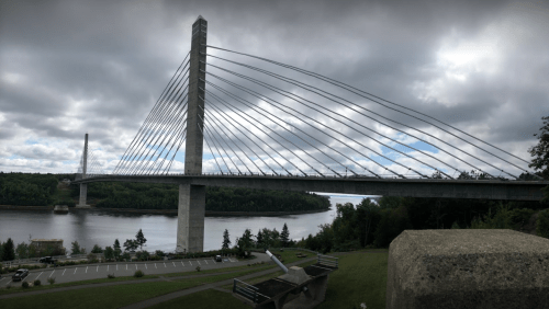 A modern cable-stayed bridge spans a river, surrounded by greenery and under a cloudy sky.
