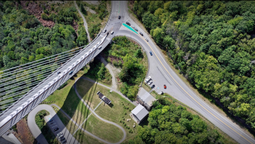 Aerial view of a bridge over a road, surrounded by lush greenery and a parking area below.