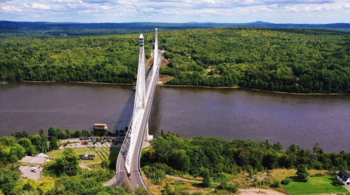 Aerial view of a tall, white suspension bridge spanning a river, surrounded by lush green forests and hills.