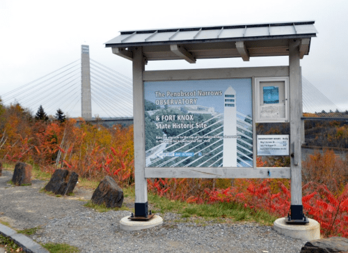Sign for the Penobscot Narrows Observatory and Fort Knox State Historic Site, with a bridge and autumn foliage in the background.