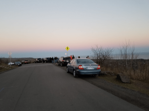Cars parked along a road at dusk, with people gathered near a scenic overlook and a pastel sky in the background.