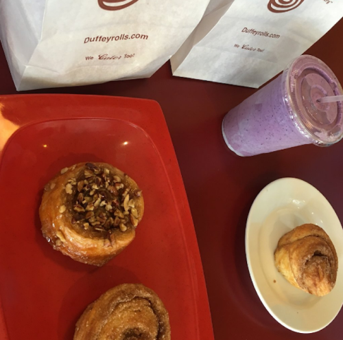 A red plate with two cinnamon rolls, a smoothie, and two paper bags from Duffeyrolls.com on a table.