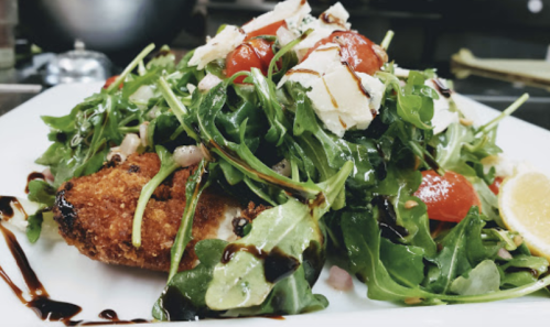 A plate of crispy chicken on a bed of arugula, cherry tomatoes, and topped with shaved cheese and balsamic drizzle.