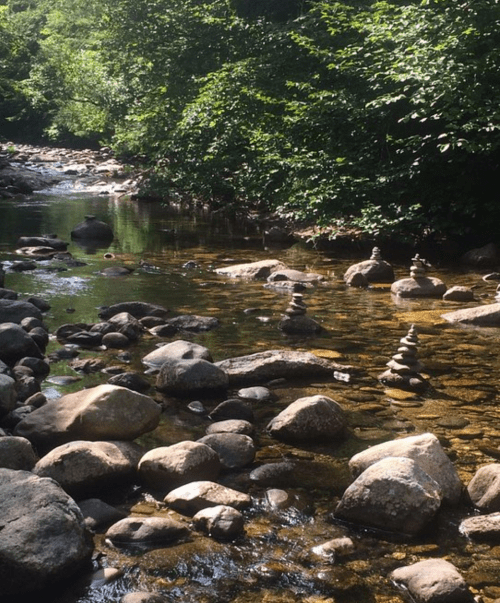 A serene stream with stacked stones along the banks, surrounded by lush greenery and smooth river rocks.