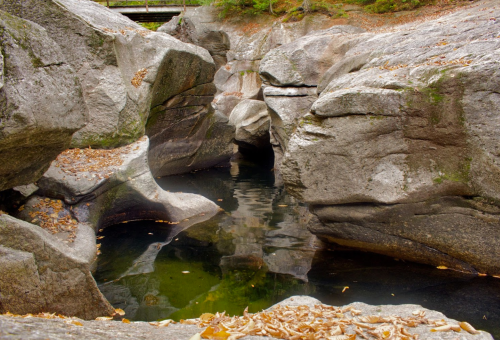 A serene rocky landscape with smooth boulders surrounding a calm, reflective pool of water. Autumn leaves scattered nearby.