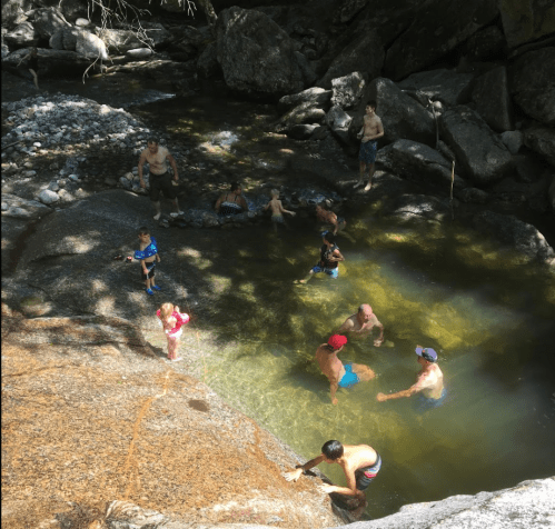 A group of people swimming and playing in a natural pool surrounded by rocks and trees on a sunny day.