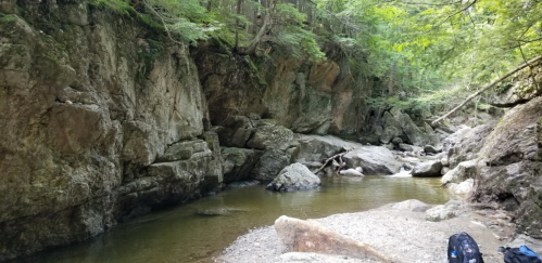 A serene rocky creek surrounded by lush greenery and steep cliffs, with calm water reflecting the trees above.