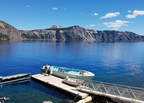A serene lake scene with boats docked at a wooden pier, surrounded by mountains and a clear blue sky.