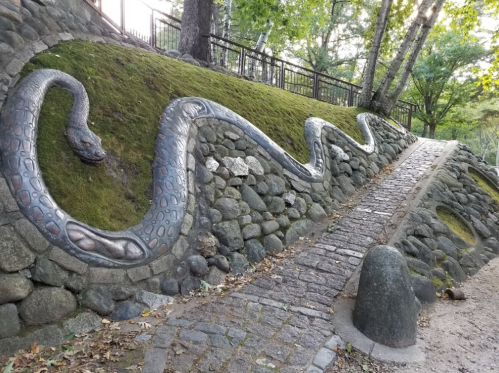 A winding snake sculpture on a moss-covered stone wall beside a pathway in a wooded area.