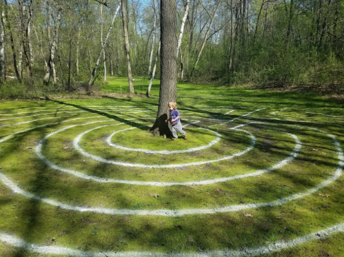 A child stands by a tree in a grassy area with a circular maze painted on the ground around them.
