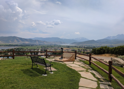 Scenic view of mountains and a valley from a park, featuring a bench and stone pathway under a partly cloudy sky.