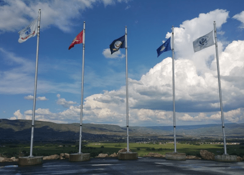 Five flags on tall poles against a backdrop of clouds and green hills, with a valley visible in the distance.
