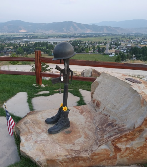 A military memorial featuring boots, a helmet, and a rifle on a rock, with a scenic view of mountains and a town.