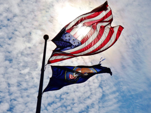 Two flags waving against a bright sky: the American flag above and the Utah state flag below.