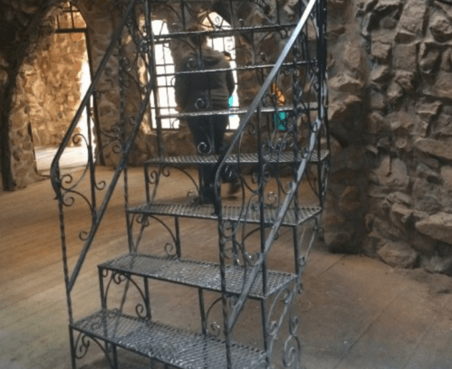 A decorative metal staircase with intricate designs, leading up in a rustic stone interior.