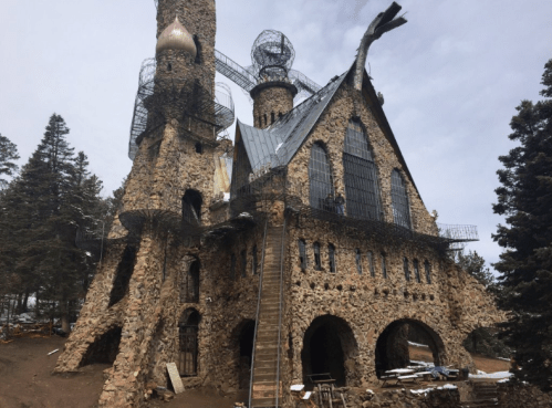 A unique stone castle with towers and a spiral staircase, surrounded by trees and under a cloudy sky.