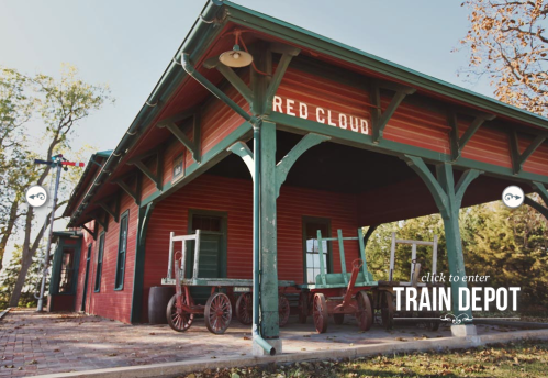 Historic red train depot with green accents, featuring wooden carts outside and a welcoming entrance.