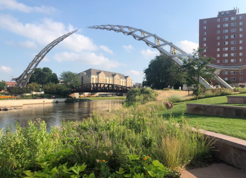 A modern arch bridge over a river, with greenery and buildings in the background under a clear blue sky.