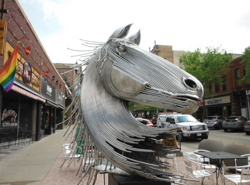 A large metallic horse sculpture made of curved metal rods, located on a city street with shops and trees in the background.
