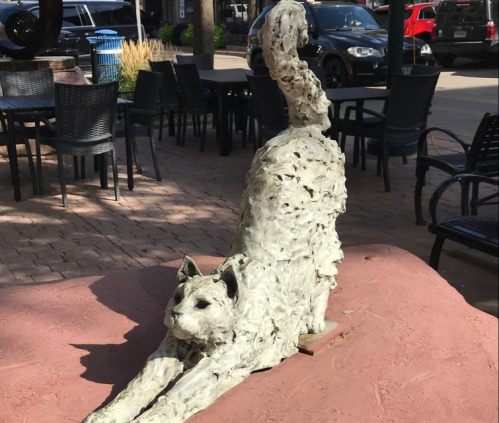 A sculpture of a stretching cat, made from textured material, displayed on a table in an outdoor setting.