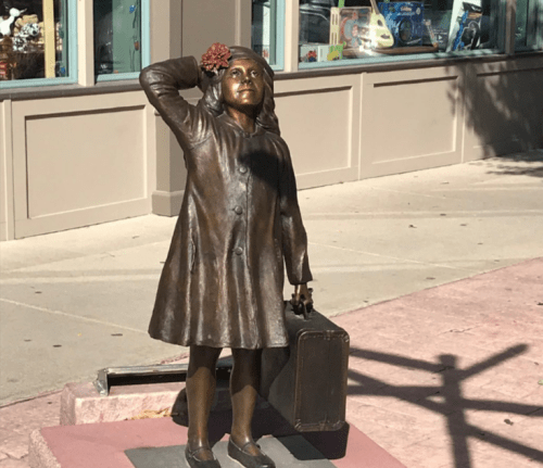A bronze statue of a girl in a coat, holding a suitcase and a flower in her hair, standing on a sidewalk.
