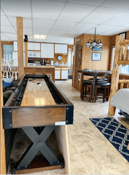 Interior view of a cozy cabin featuring a shuffleboard table, dining area, and kitchen in a rustic setting.