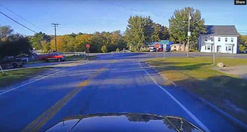 A view of a rural road intersection with a stop sign, trees, and a few buildings in the background.
