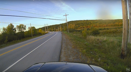 A view of a rural road lined with power lines, surrounded by trees with autumn foliage and a distant hill.