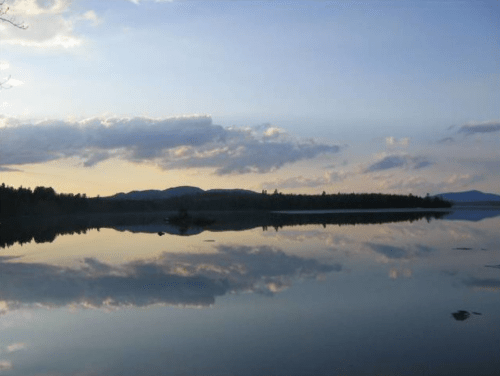 A serene lake at sunset, reflecting clouds and mountains in calm waters, surrounded by trees.