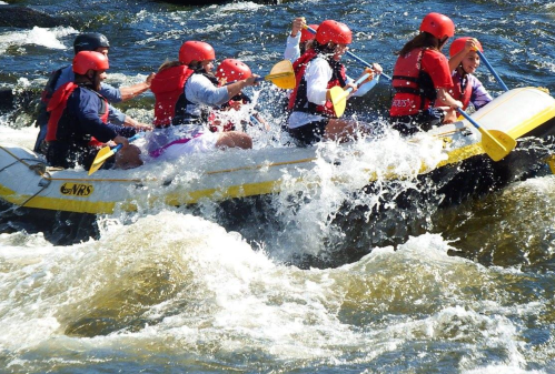 A group of people in red helmets and life jackets rafting through turbulent waters, splashing water around them.