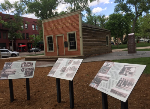 Historic log cabin with informational plaques in a park, surrounded by trees and nearby buildings.