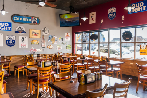 A cozy restaurant interior with wooden tables, chairs, and various beer signs on the walls, featuring a large window.